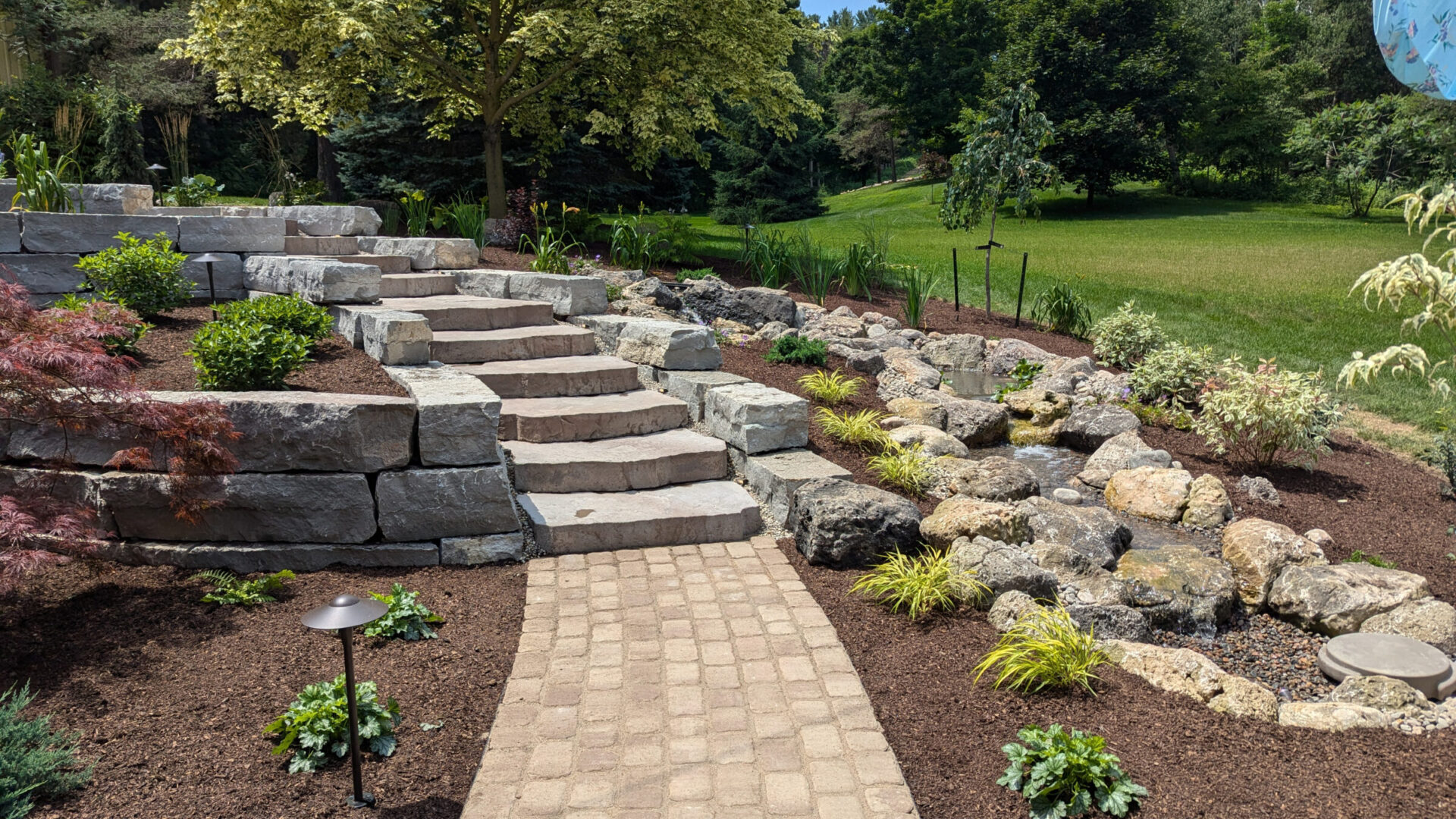 A beautifully landscaped garden with stone steps, lush greenery, decorative rocks, and a small stream, creating a peaceful, natural setting.