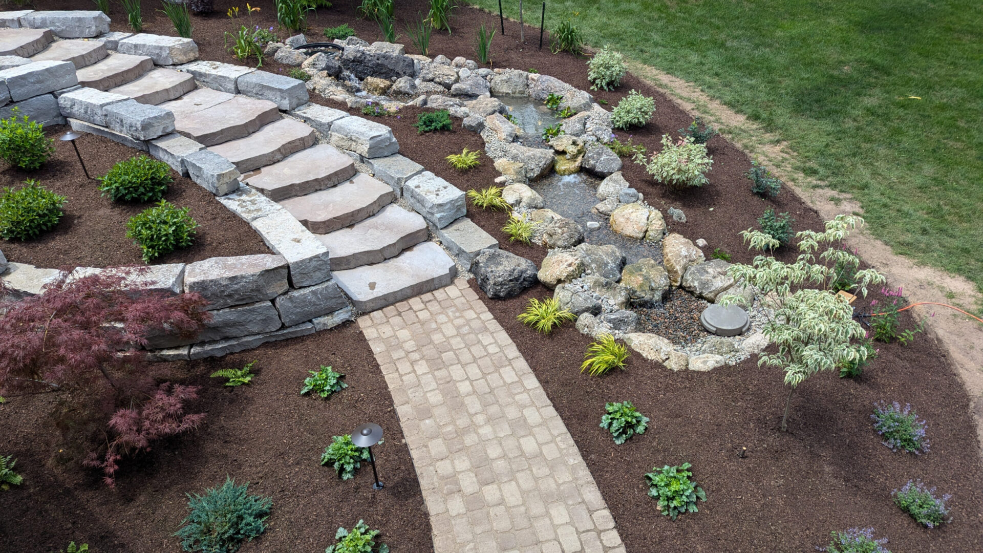 A landscaped garden featuring stone stairs, a rock-lined water feature, and lush greenery in a serene outdoor setting.