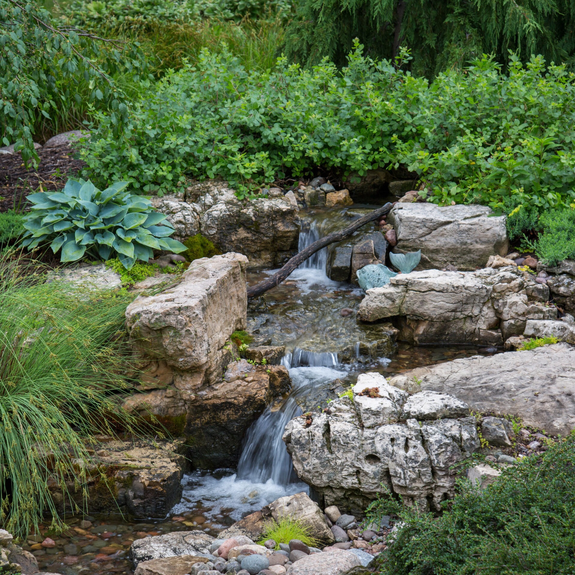 A serene garden scene featuring a small waterfall cascading over rocks, surrounded by lush greenery and vibrant plants.