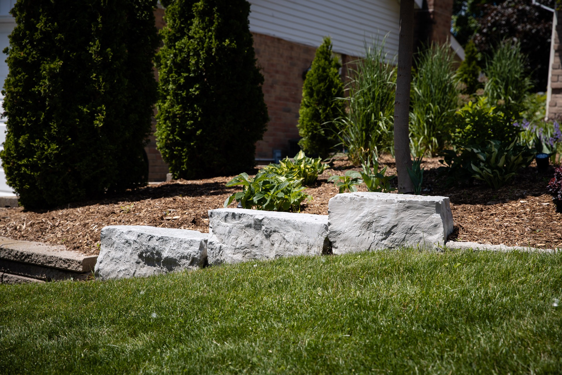 A well-maintained garden with rocks, green plants, and mulch, bordered by neatly trimmed grass, suburban house visible behind.