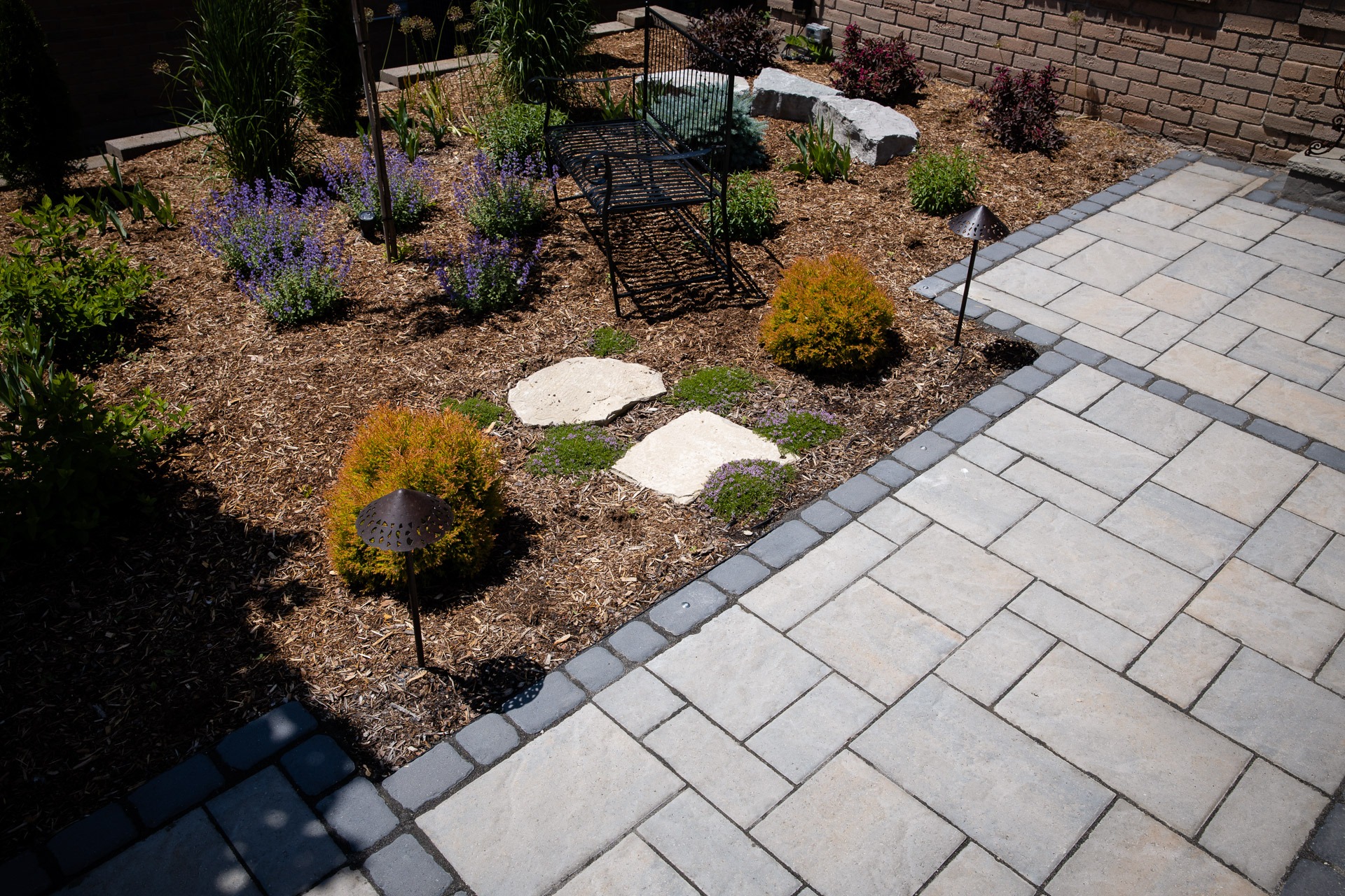 Garden with a stone pathway, metal bench, and various plants surrounded by mulch. Adjacent is a paved area with brick pattern.