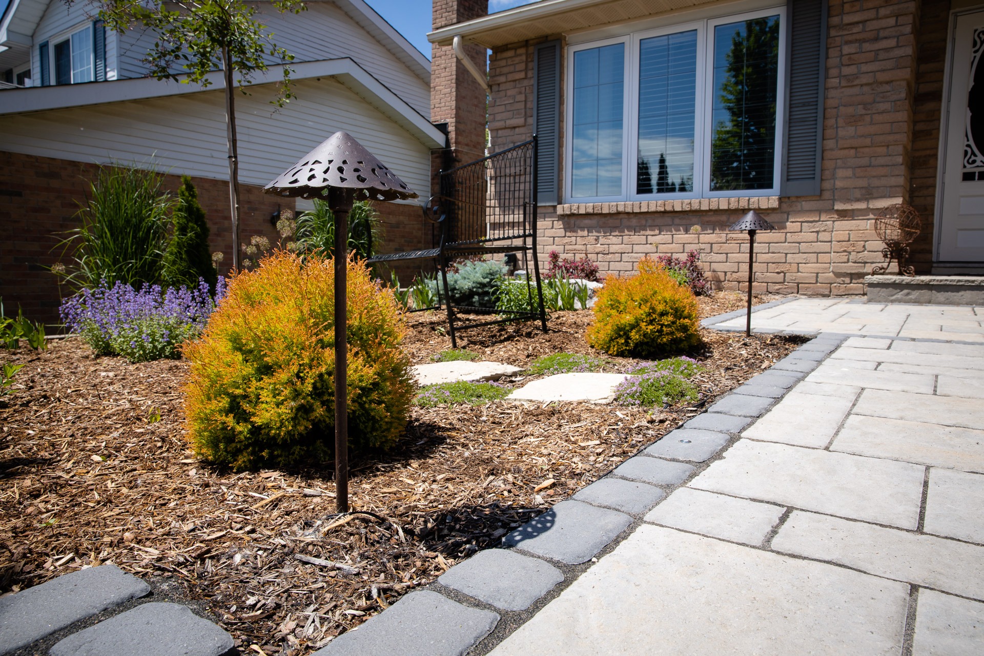 A landscaped garden with mulch, shrubs, pathway lights, and stepping stones leads to a brick house with large windows.