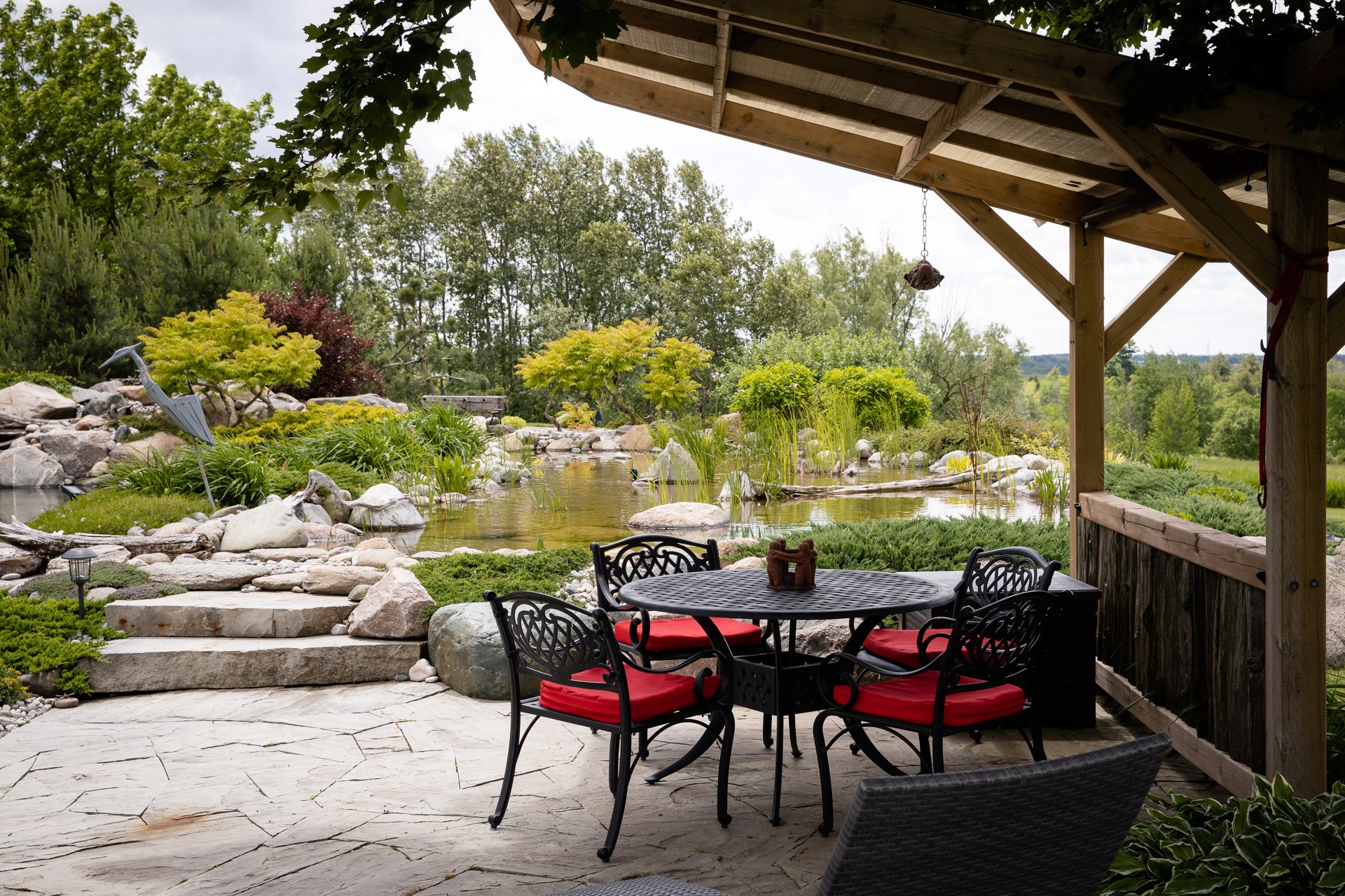 A serene backyard patio with a table and chairs overlooks a pond, surrounded by lush greenery and stone landscaping under a wooden pergola.