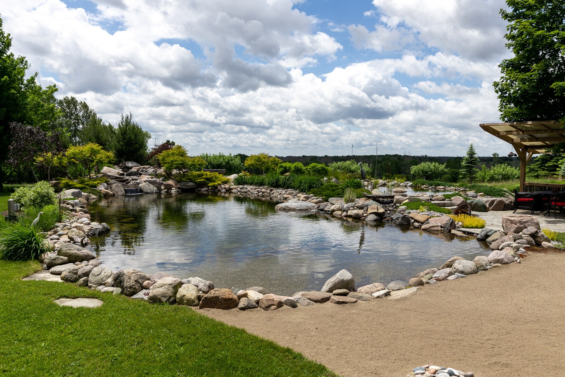 A tranquil garden scene with a stone-bordered pond, lush greenery, and a wooden pergola under a partly cloudy sky. No people present.