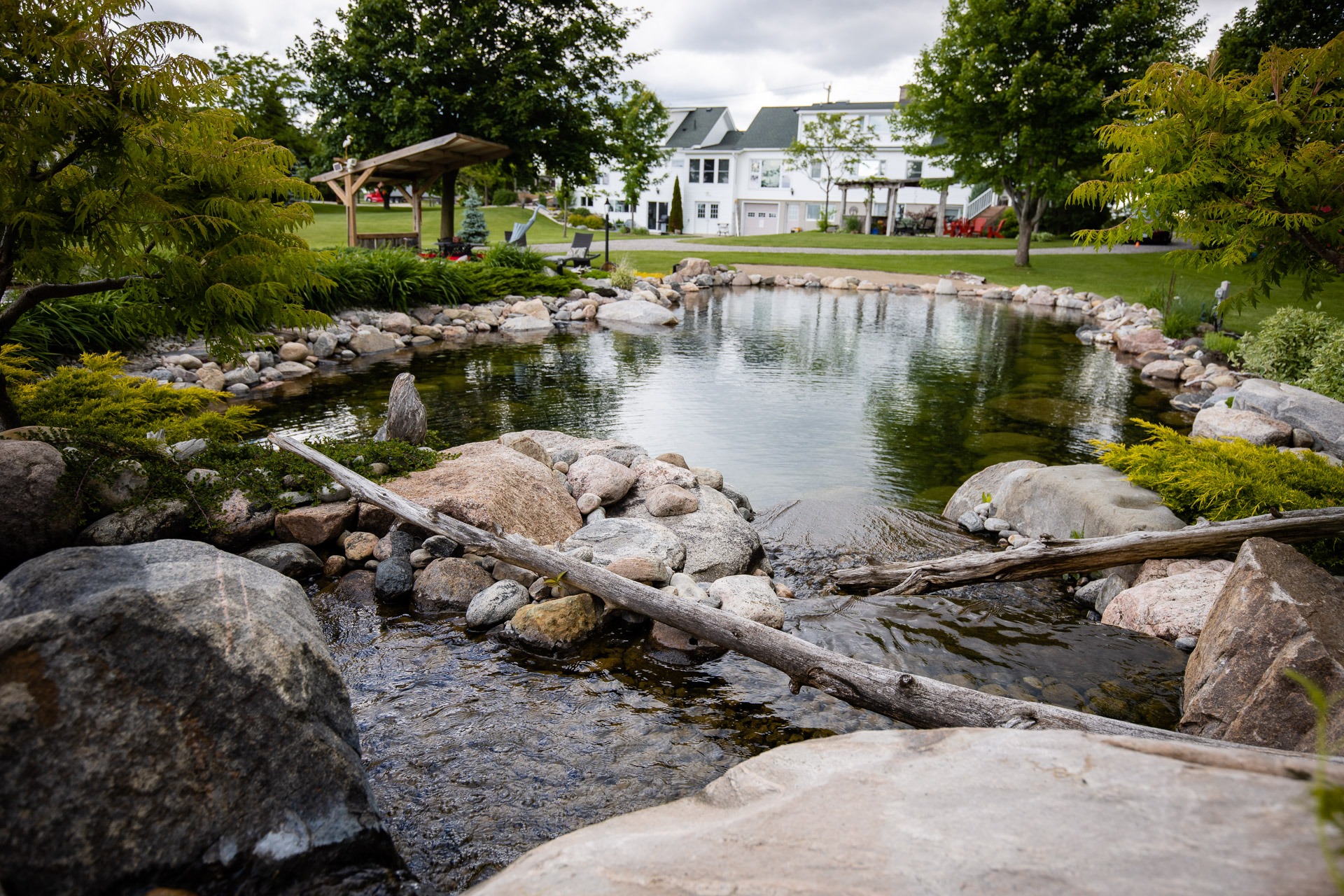 A tranquil garden pond surrounded by stones and greenery, with a white house and pavilion in the background under a cloudy sky.