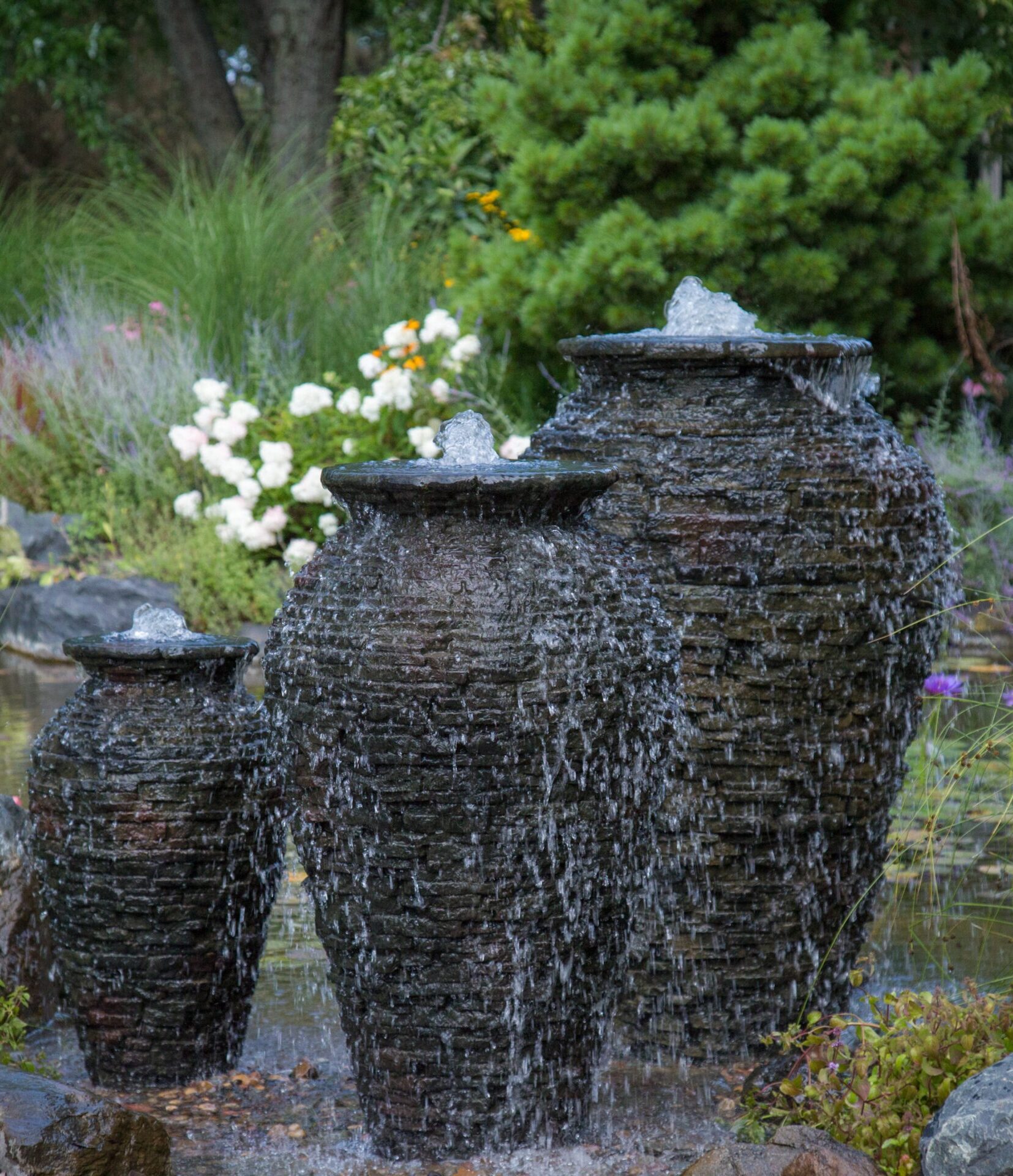 Three stone fountains with cascading water surrounded by lush greenery and vibrant flowers, creating a serene garden atmosphere.