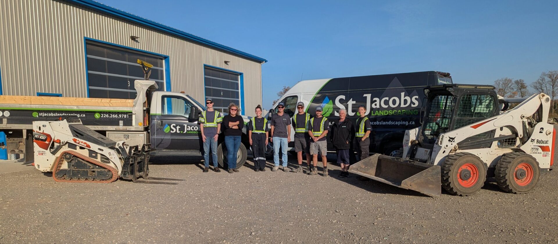Seven people stand in front of landscaping vehicles and equipment outside a metal building, wearing work apparel and smiling at the camera.