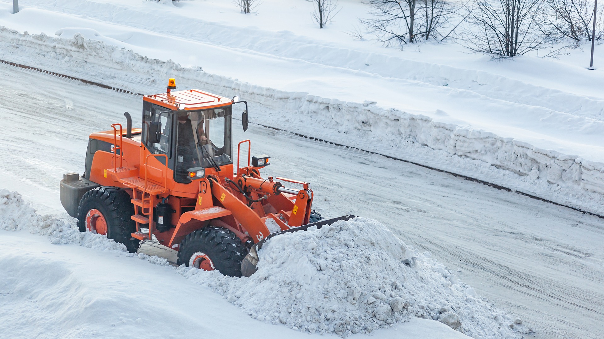 An orange snowplow clears snow on a wide road. The area is covered in heavy snow with trees lining the background.