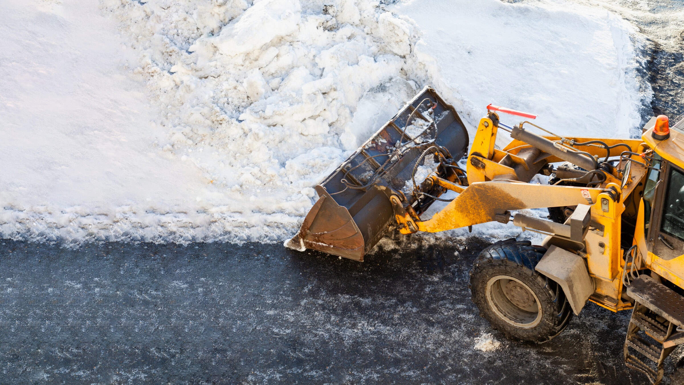 A yellow snowplow removes snow from a road, clearing a snowy area. Bright sunlight casts shadows, highlighting winter maintenance work in process.