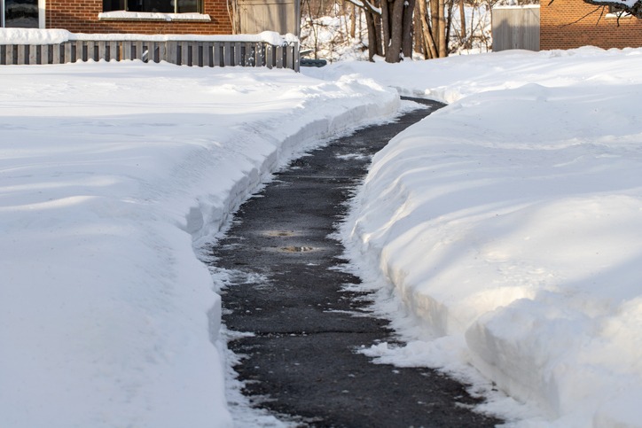 A snow-covered path leading between snowbanks, with a brick building and trees in the background, under a clear sky.