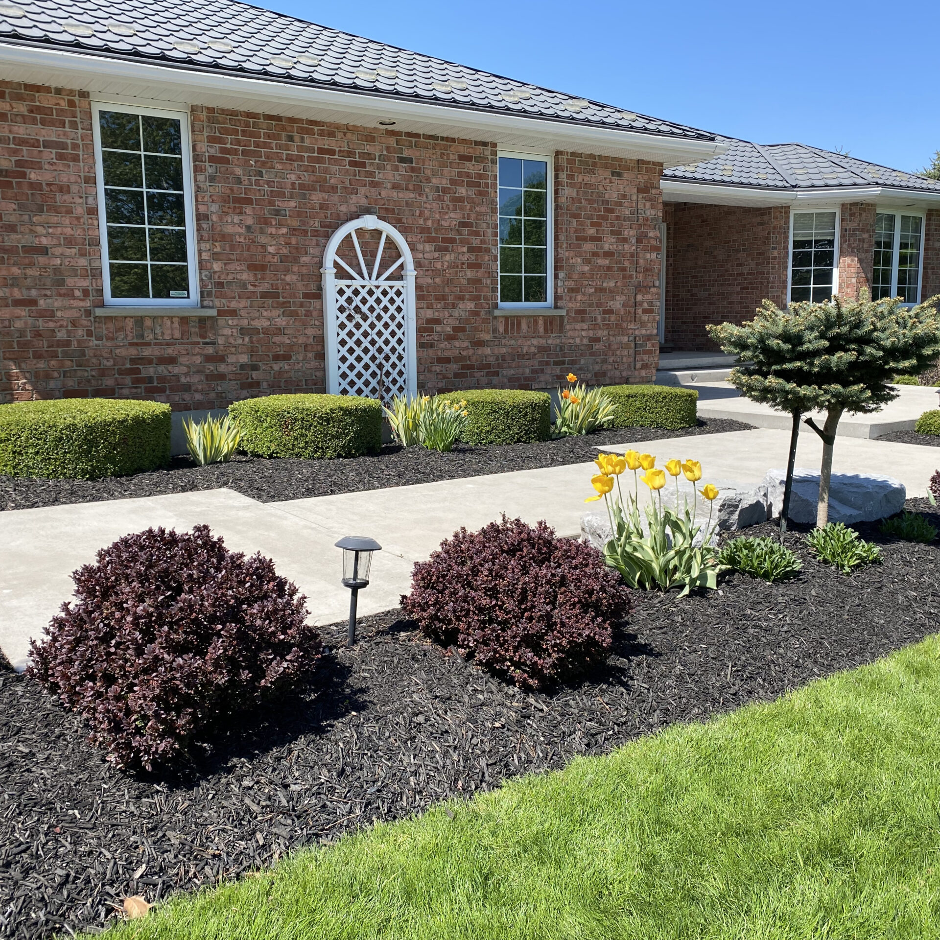 Brick house with neatly landscaped garden, featuring hedges, colorful flowers, small tree, and a pathway. Clear blue sky in background.