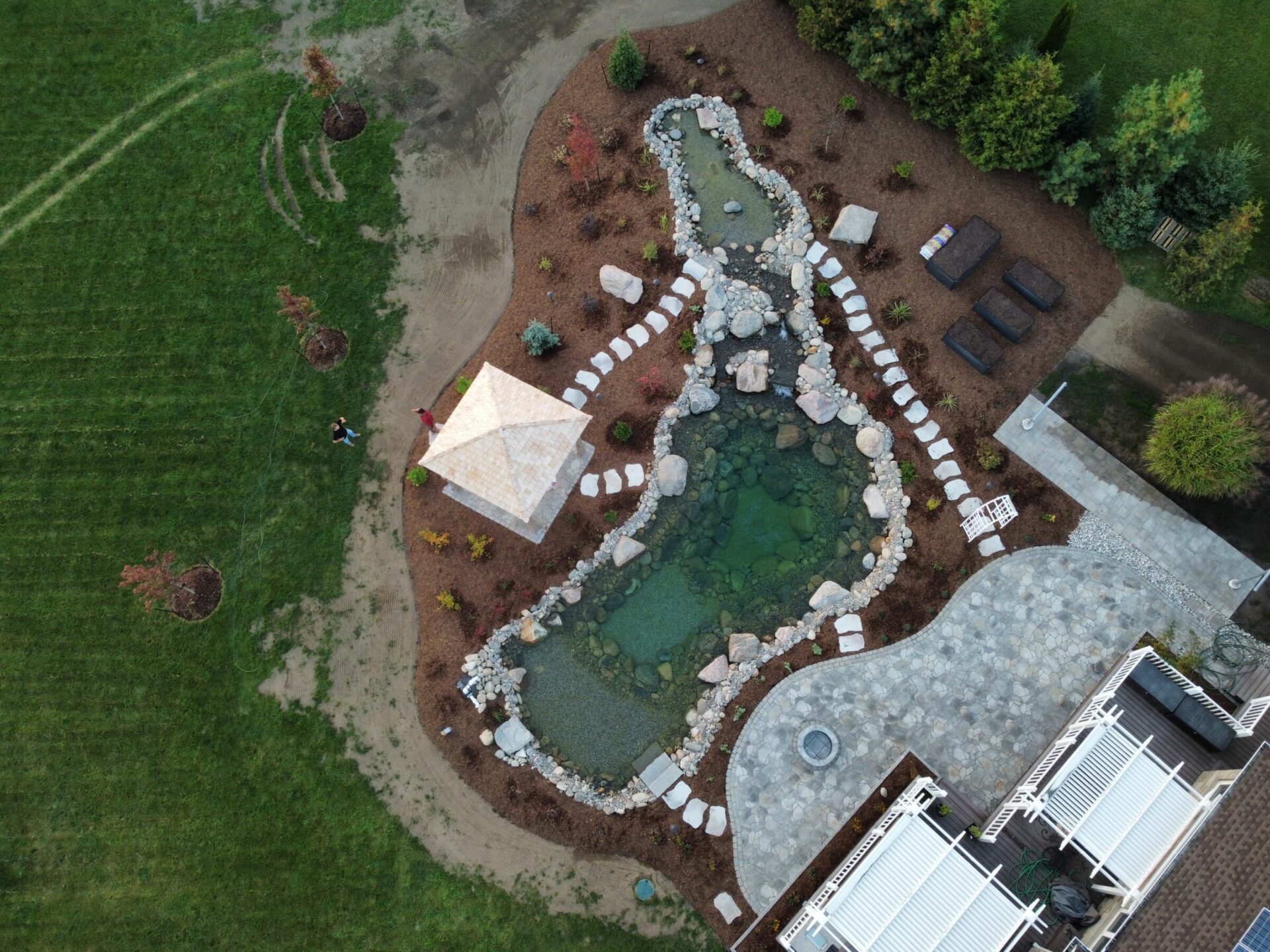 Aerial view of a landscaped garden with a pond, stone pathways, gazebo, grassy area, and two people walking nearby.