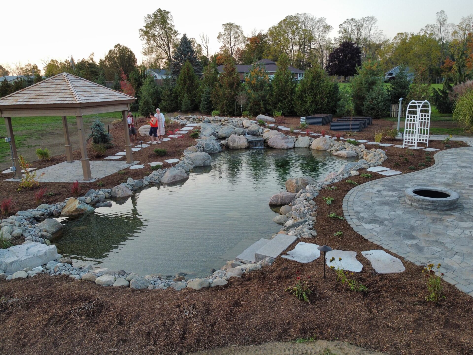 A landscaped garden with a pond, gazebo, stone pathway, and three people. Surrounding trees and greenery create a serene outdoor setting.