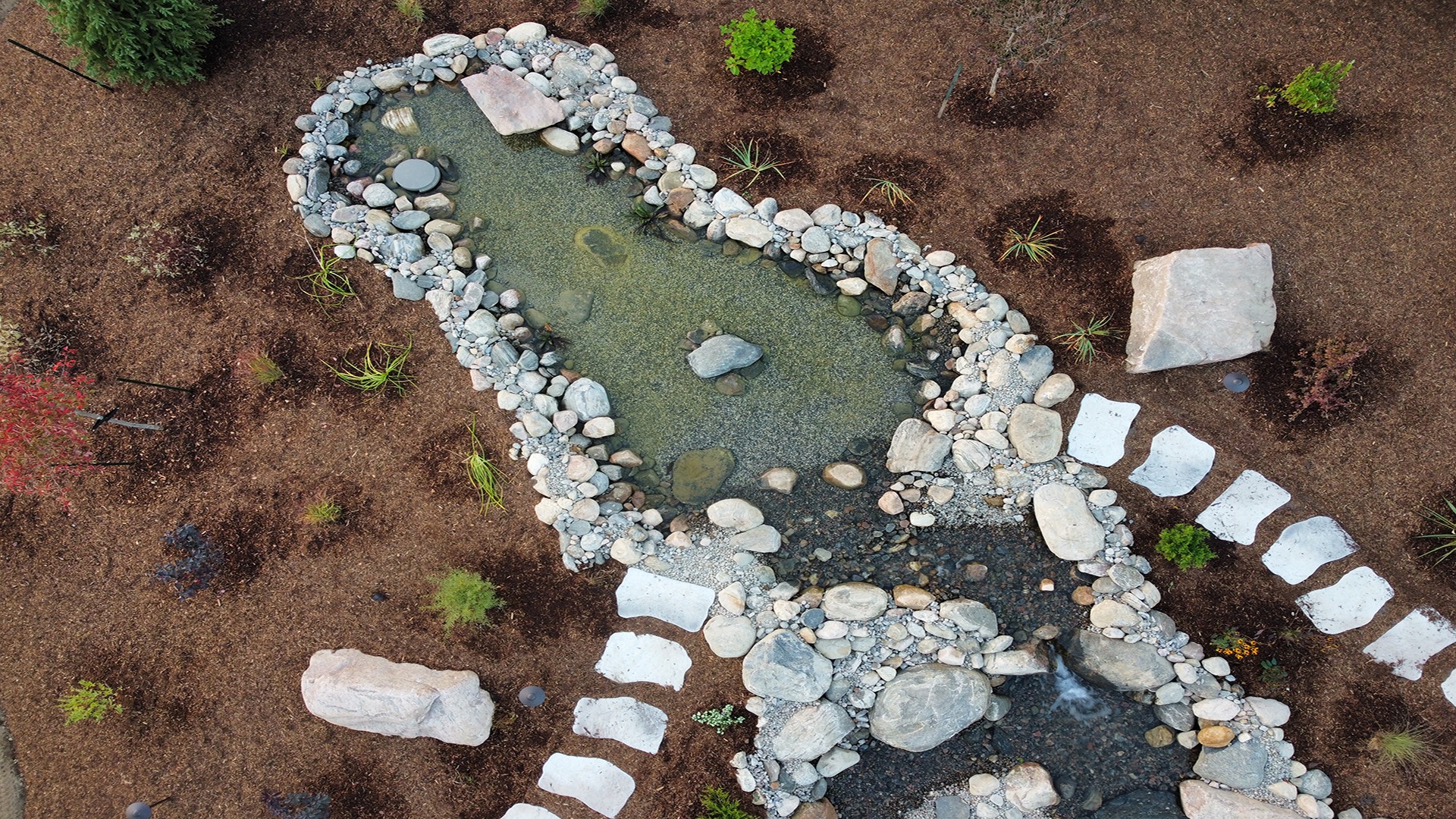 Aerial view of a stone-lined garden pond, surrounded by mulch, stepping stones, and scattered plants in a landscaped outdoor setting.