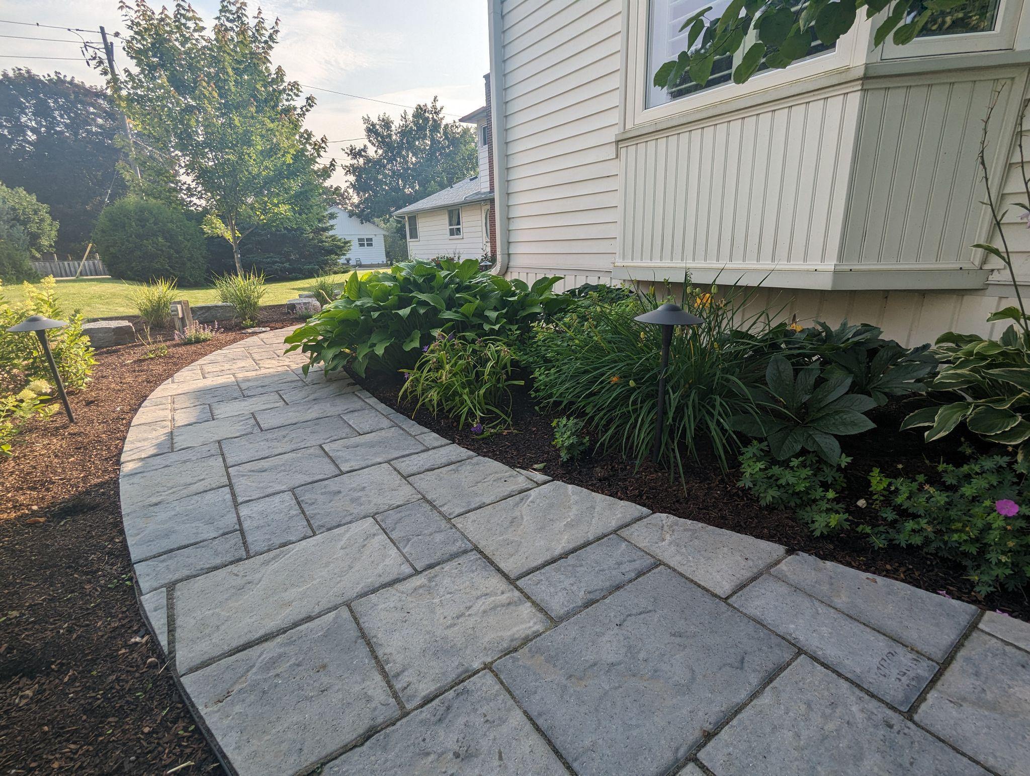 Front yard hardscape renovation featuring a cobblestone-style paver walkway and raised garden bed beside a brick home. Fresh mulch, small ornamental trees, and clean edging create a neat and polished curb appeal.