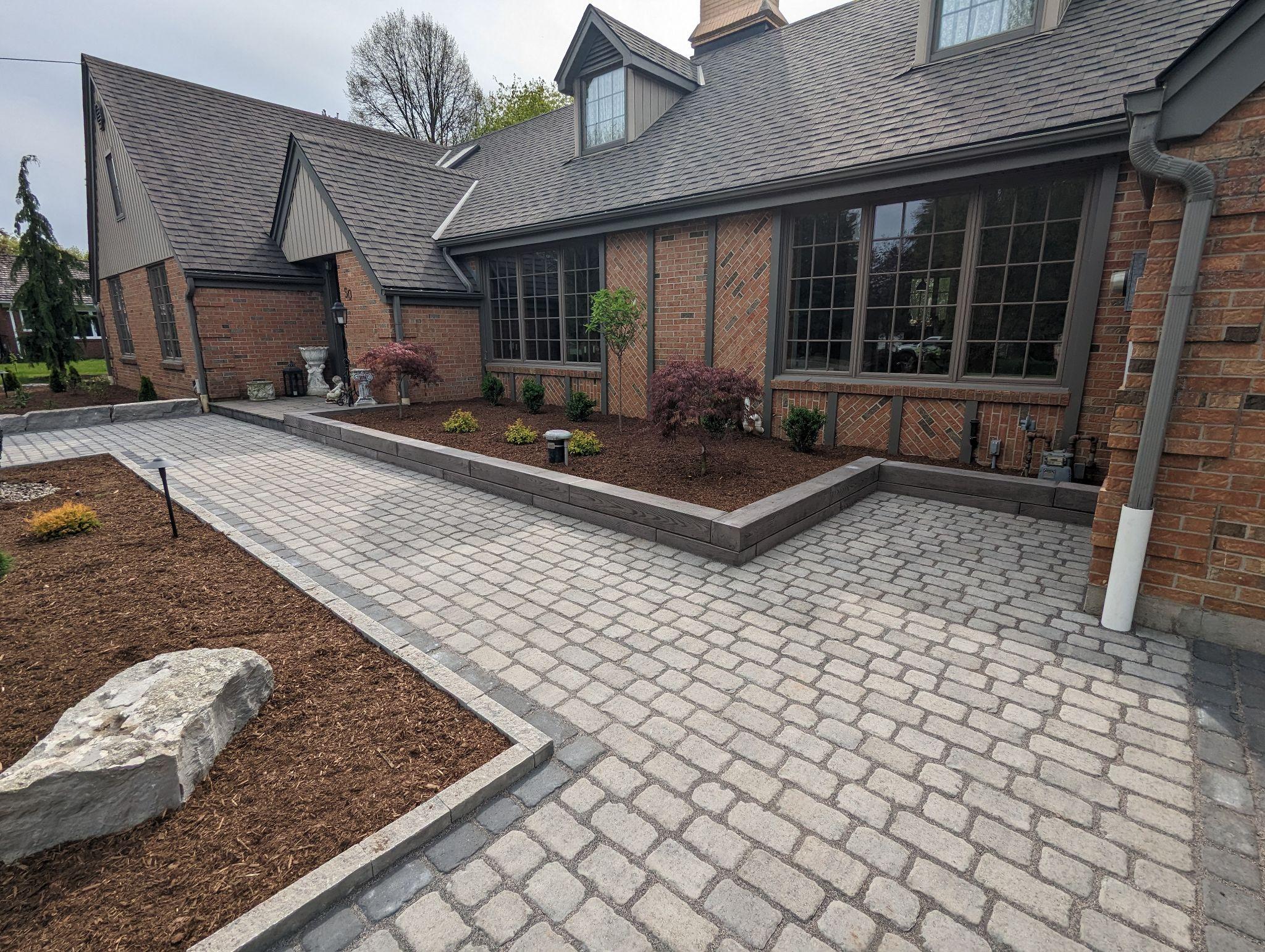 Curved stone walkway bordered by garden beds with hostas, ornamental grasses, and mulch beside a light-coloured house. Landscape lighting and mature trees provide a welcoming, natural look.