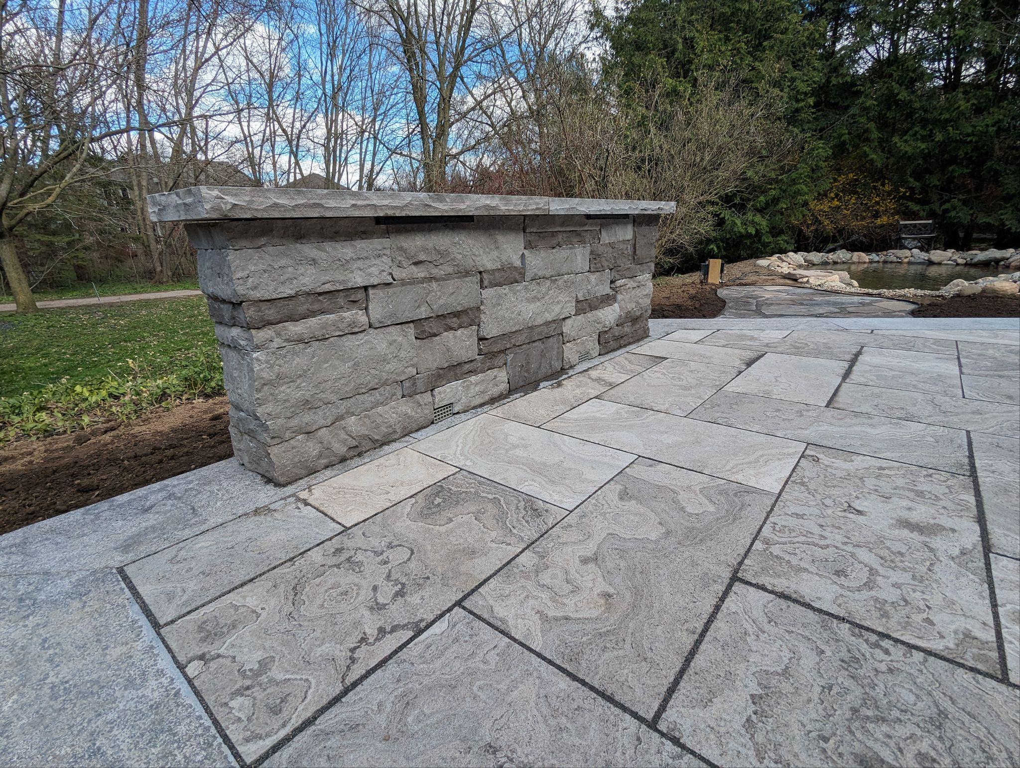 Freestanding stone bar on a large natural‑stone patio, with a pond and path in the background.