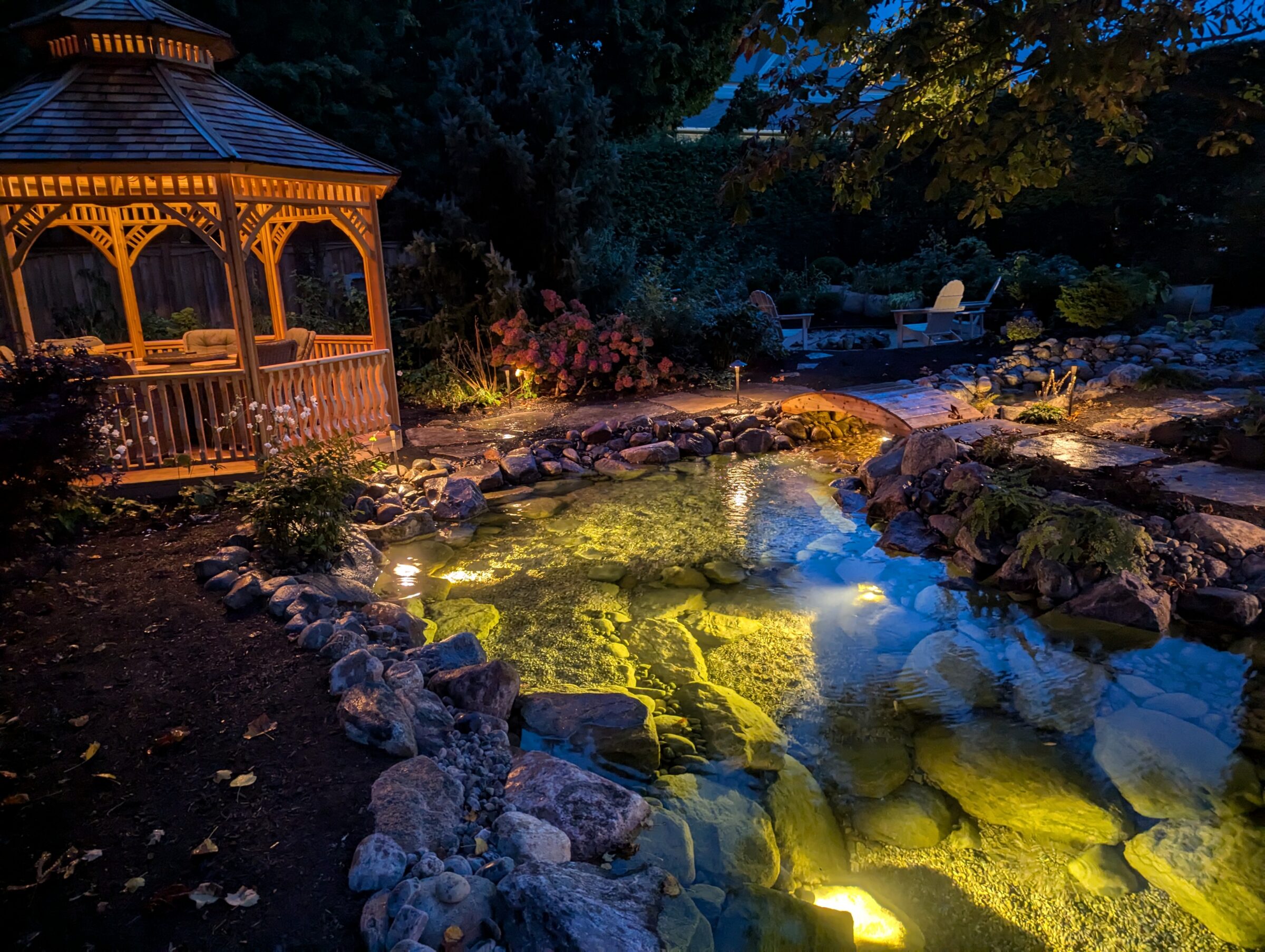 Lit wooden gazebo beside a rock-lined pond and footbridge at dusk