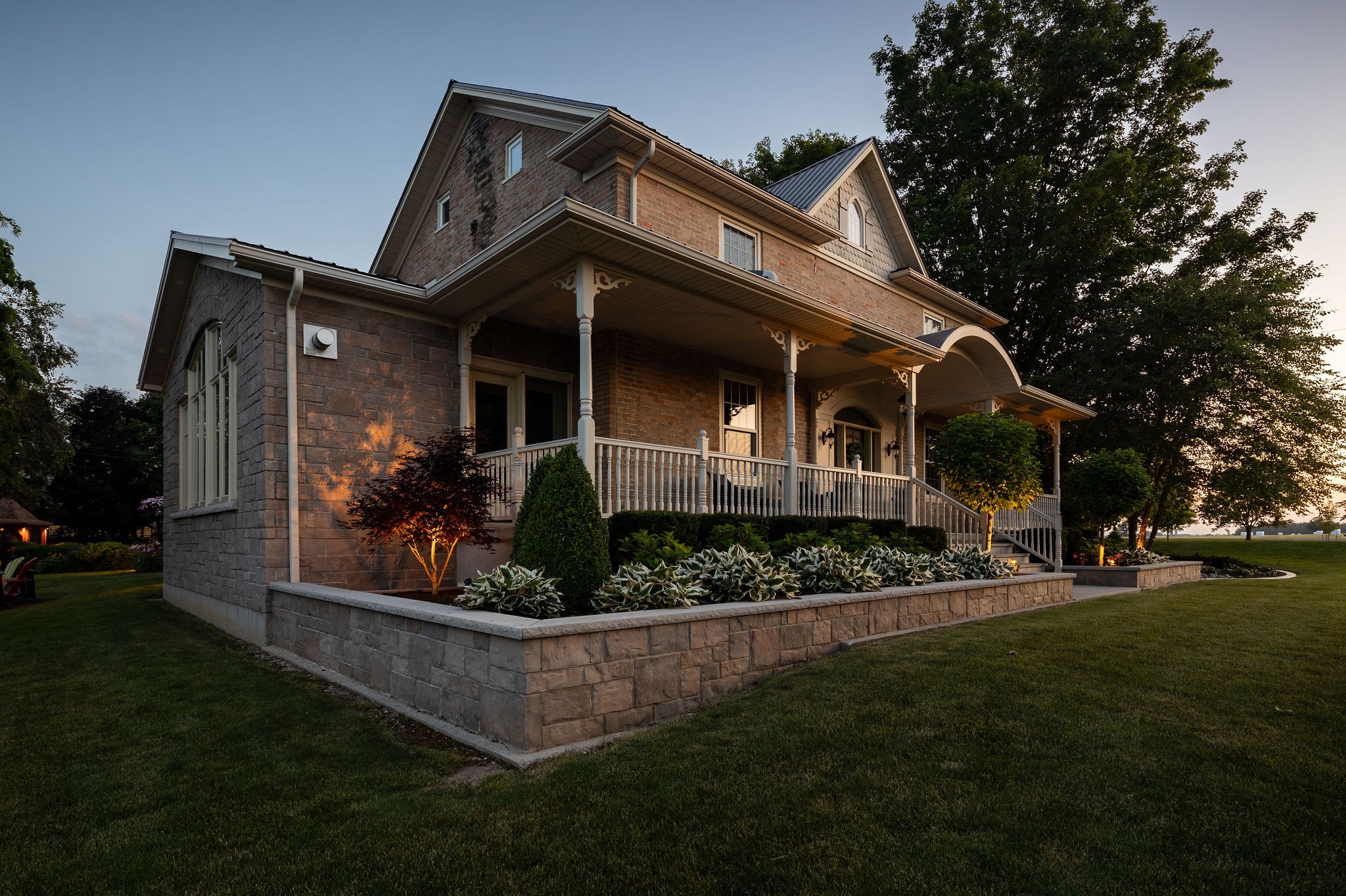 Two-story brick home with wraparound covered porch, stone retaining wall, and dusk landscape lighting