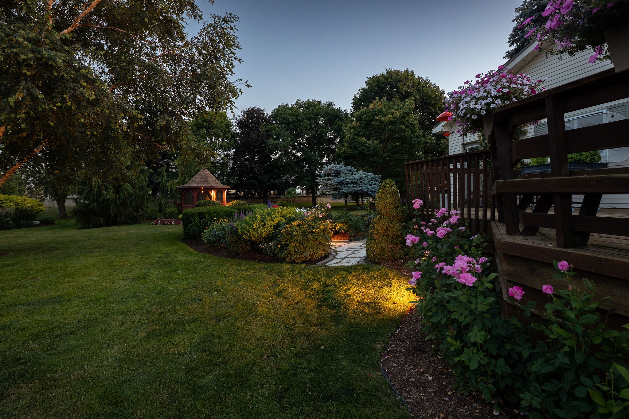 Backyard lawn and garden path leading to a lit gazebo at dusk, deck with hanging flowers in foreground