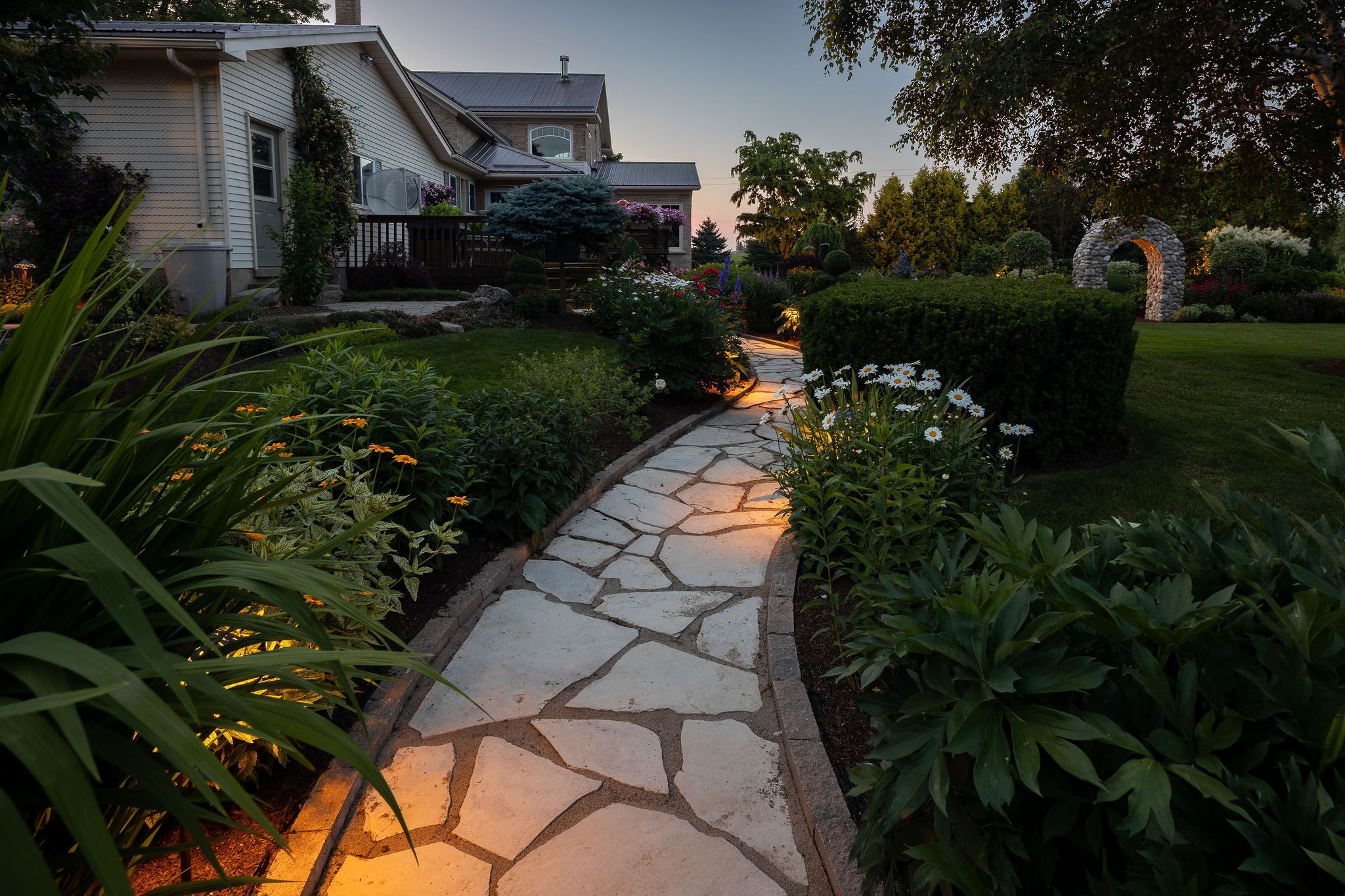 Curved stone garden pathway lit at dusk leading toward a house and stone archway