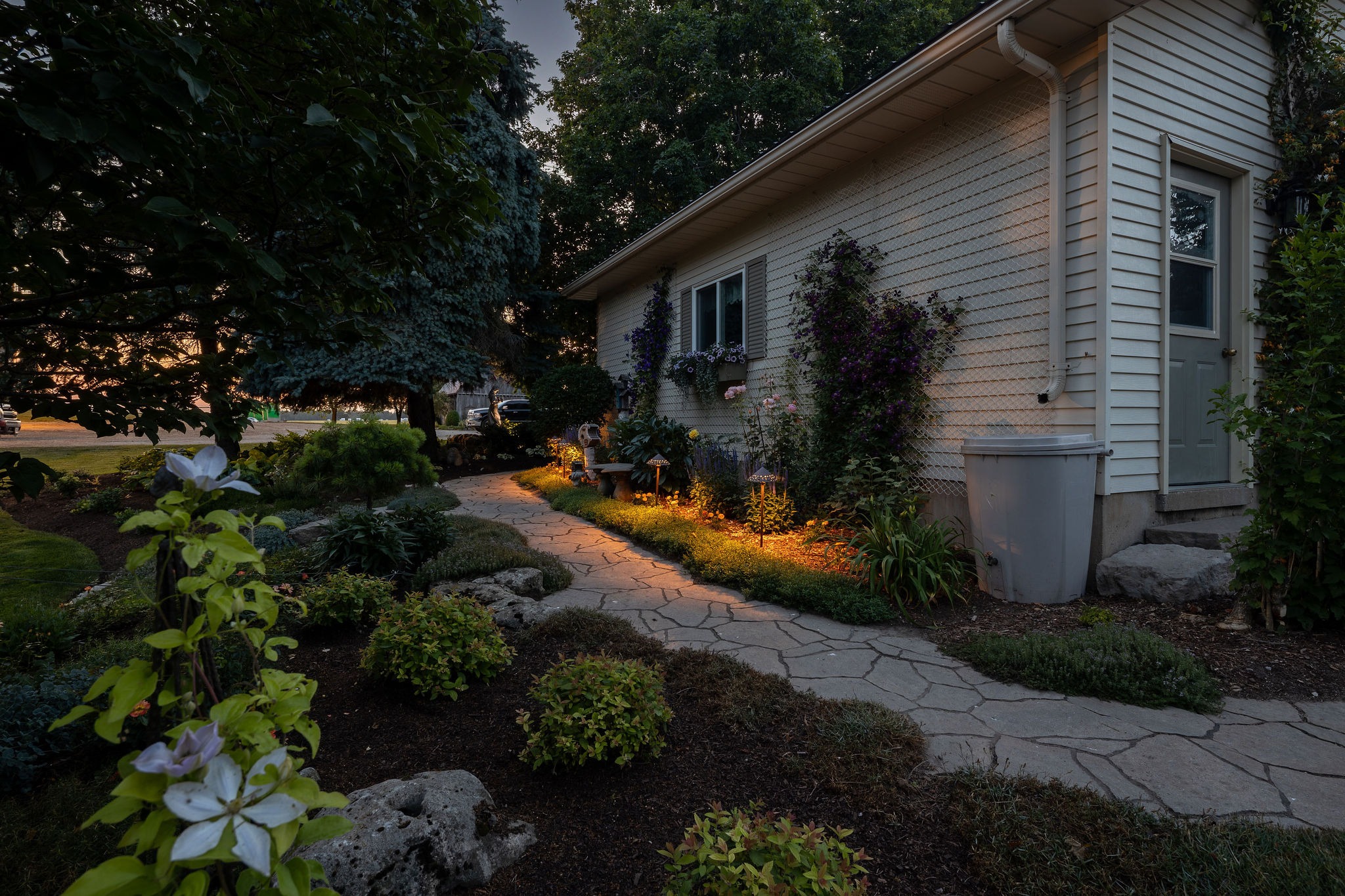 Stone walkway lit by landscape lighting along a flower bed beside a house