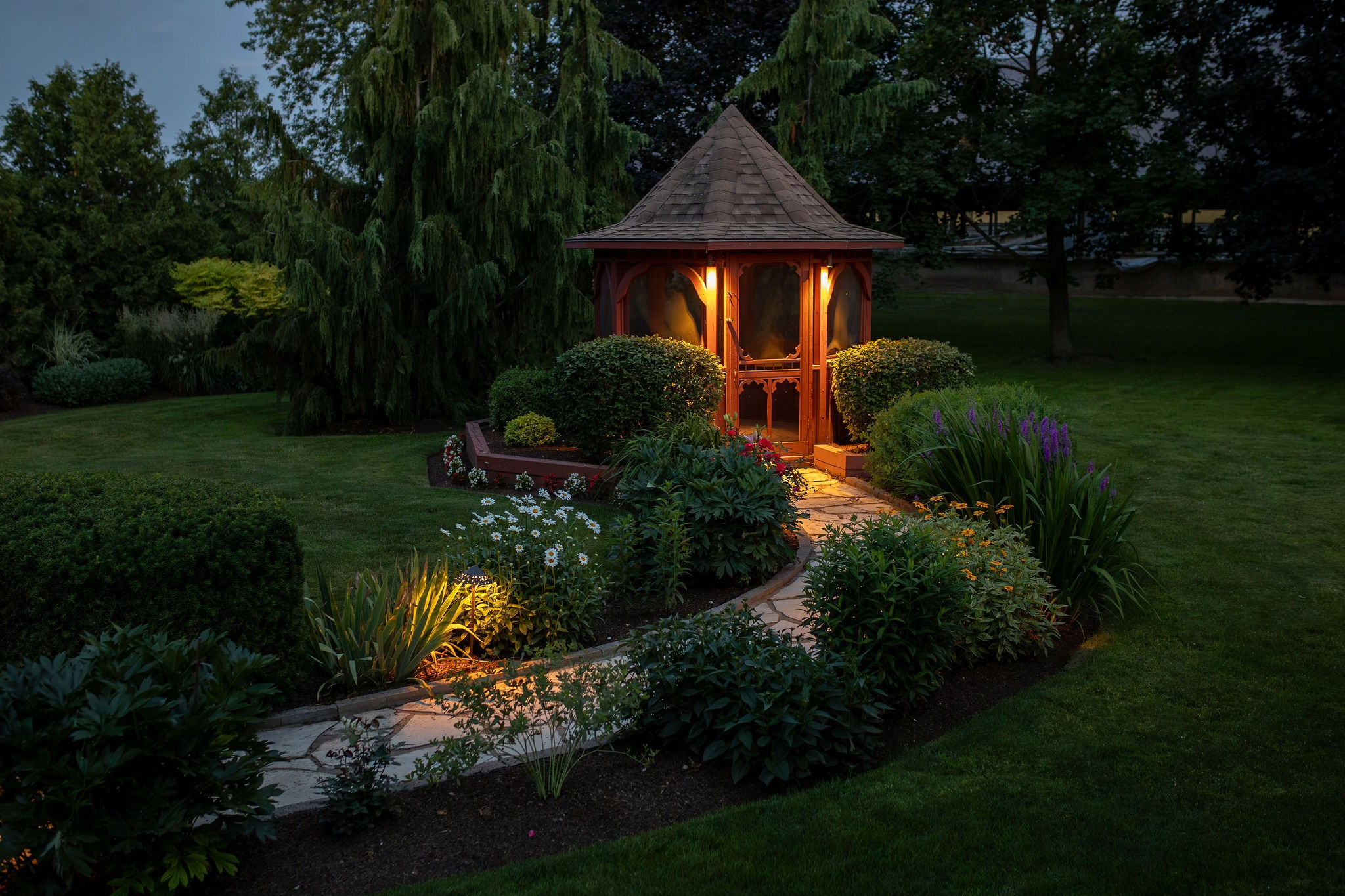 Illuminated garden gazebo with curved stone pathway through flower beds at dusk